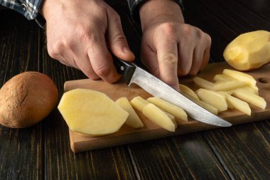 Bir insan, tahta kesme tahtasındaki patatesleri ince parçalara ayırmak için bıçak kullanıyor. Sahne, doğal ışık eşliğinde bir mutfakta geçiyor..