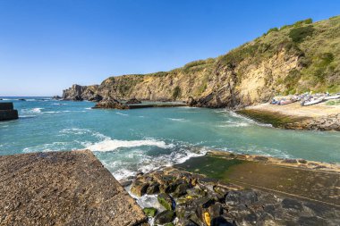 Portekiz 'deki Azenha do Mar' ın kıyı manzarasında turkuaz sular, kayalık kayalıklar ve berrak mavi gökyüzü altında küçük bir balıkçı limanı yer alıyor.