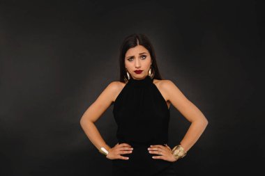 Tatmin olmayan, uzun siyah saçlı, parlak makyajlı, altın küpeli, siyah arka planda kameraya bakan ciddi bir kız. Yüksek kalite fotoğraf