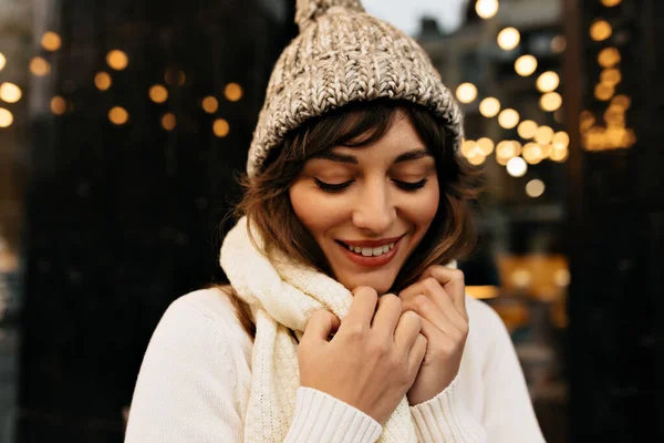 Incredible charming lady in knitted white hat and knitted sweater smiling on background of Christmas lights. High quality photo