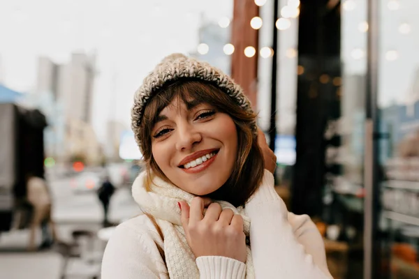 Blissful girl in stylish winter outfit posing in outdoor cafe with modern exterior. Photo of merry caucasian woman enjoying Holiday. High quality photo