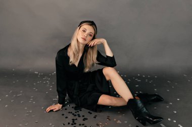 Stüdyoda, siyah elbise ve çizme giyen güzel sarışın bir kadının gri arka planda konfeti ile poz verdiği bir fotoğraf. Yüksek kalite fotoğraf