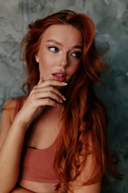 Uzun dalgalı kızıl saçlı, güzel bir kadının portresi. Çenesine dokunuyor ve gri arkaplana bakıyor. 