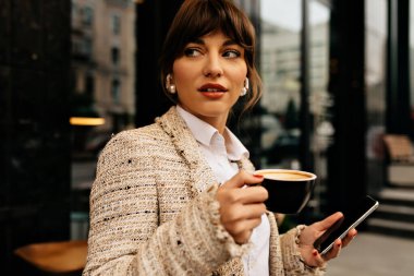 Şık, şık bir iş kadını, beyaz elbiseler giymiş, kahve içerken şehir arka planında akıllı telefon kullanıyor. Yüksek kalite fotoğraf