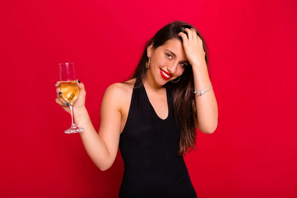Pretty happy woman straightens hair with a glass of champagne wearing black clothes over red background. High quality photo