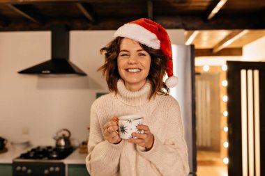 Kapalı alanda, Noel Baba şapkası takan ve elinde Noel ışıkları bardağıyla oturan dalgalı saçlı kadının portresini kapat. Yüksek kalite fotoğraf