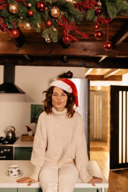 Kıvırcık saçlı, örgü elbiseli ve Noel Baba şapkalı sevimli bir kadın mutfakta oturuyor ve Noel partisini bekliyor. Yüksek kalite fotoğraf