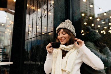Örgü beyaz şapkalı ve fularlı, şehir kafeteryasının arka planında kahve molası veren çok sevimli bir kız. Yüksek kalite fotoğraf