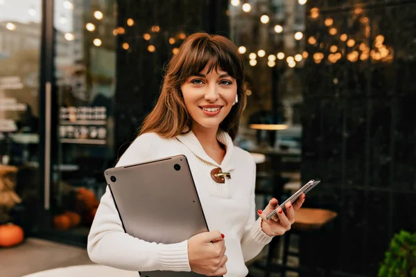 Successful charming lady with laptop and smartphone posing outside in city lights . High quality photo