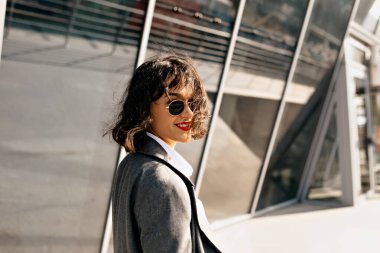 Dışarıda, kısa dalgalı saçlı, gri ceketli ve güneş ışığında iş yerinin yakınında yürüyen gözlüklü hoş bir kadının portresi var. Yüksek kalite fotoğraf