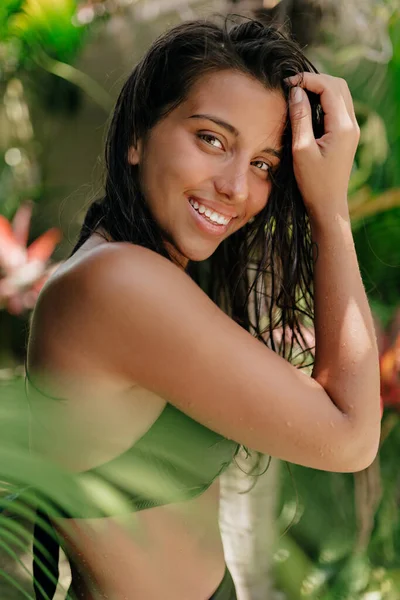 Außenporträt einer attraktiven jungen, braun gebrannten Frau mit dunklem, nassem Haar, die lächelt und vor der Kamera vor dem Hintergrund exotischer Pflanzen posiert — Stockfoto