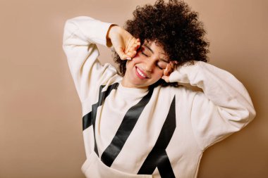 Kıvırcık saçları olan, beyaz kazak giyen, gözleri kapalı gülümseyen ve izole arka planda uzanan rahat ve çekici bir kız. Yüksek kalite fotoğraf