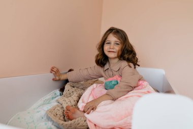 Dalgalı saçlı heyecanlı çocuk yatakta eğleniyor, pijama giyiyor ve oynuyor. Yüksek kalite fotoğraf