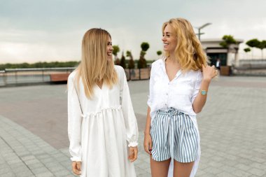 Two charming women with blond hairstyles wearing while summer clothes walking down the street in summer day