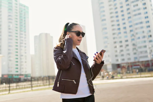 Wanita bergaya menarik dengan jaket kulit dan t-shirt putih dengan kacamata hitam mendengarkan musik ketika berjalan di kota di bawah sinar matahari — Stok Foto