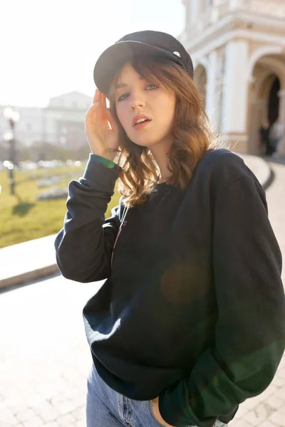 Стильная симпатичная дама в кепке позирует перед камерой во время прогулки по солнечному городу — стоковое фото