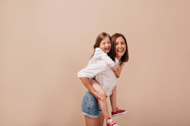 Beyaz tişörtlü ve şortlu mutlu bir kadın kızını sırtında tutuyor ve bej arka planda kameraya gülümsüyor. Yüksek kalite fotoğraf