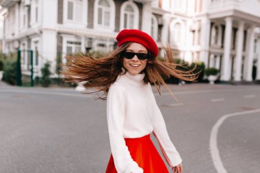 Heyecanlı, mutlu, uzun saçlı, kırmızı bereli ve etekli bir kız şehirde yürürken kameraya doğru dönüyor. Yüksek kalite fotoğraf