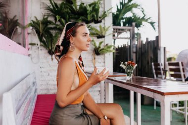 Güzel bir kızın profil portresi yaz kıyafetleri içinde şık bir açık hava restoranında oturuyor ve arka planda egzotik bitkilerin kahvesiyle oturuyor. Yüksek kalite fotoğraf