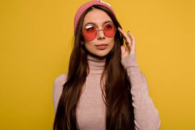 Pembe şapkalı, pembe gözlüklü, uzun saçlı, şık bir kadın sarı arka planda poz veriyor. 