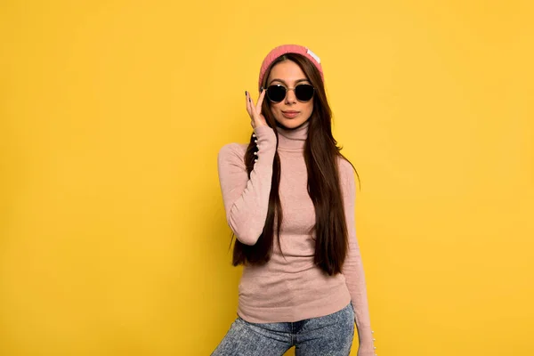 Elegante chica europea con el pelo largo y oscuro usando gafas de sol negras tocando sus gafas y posando ante la pared amarilla —  Fotos de Stock