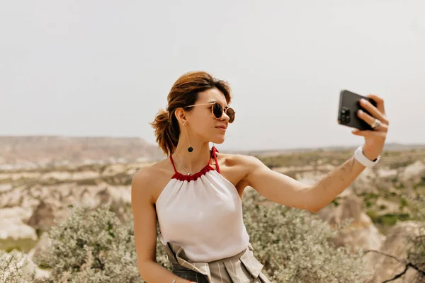 Gambar wanita cantik yang tersenyum di luar ruangan dengan pakaian musim panas membuat selfie di latar belakang pegunungan di bawah sinar matahari — Stok Foto