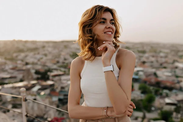 Романтичная красивая девушка с вьющимися волосами в белой футболке, смотрящая в сторону и улыбаясь на фоне городского пейзажа — стоковое фото