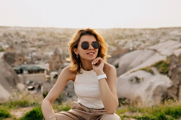 Gadis cantik romantis dengan rambut pendek dengan kacamata hitam tersenyum dan memandang jauh di bawah sinar matahari di antara gunung-gunung dengan pandangan di kota — Stok Foto