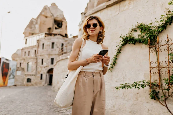 Gadis cantik bergaya dengan gaya rambut pendek berjalan dengan smartphone di antara kota tua di bawah sinar matahari — Stok Foto