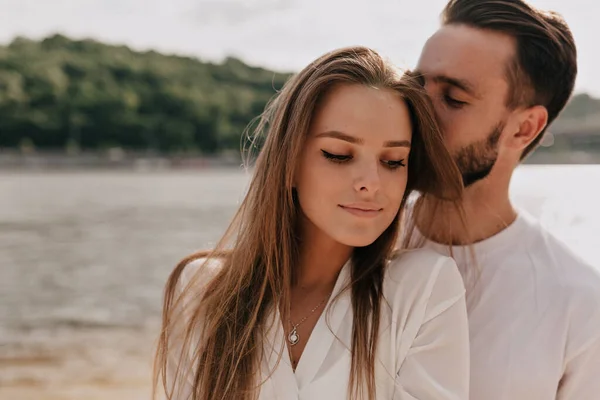 Sensitive pretty girl with long light-brown hair smiling while her boyfriend is kissing her on the sandy beach. High quality photo