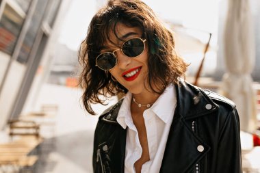 Kırmızı dudaklı ve dalgalı saçlı hoş bir kadın. Deri ceketli çekici bayan kameraya gülümsüyor. Yüksek kalite fotoğraf