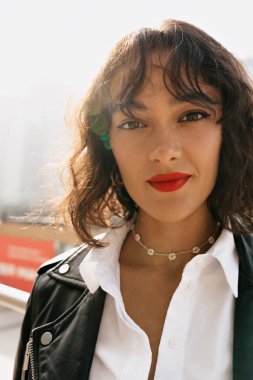 Neşeli siyah saçlı, kırmızı dudaklı, modaya uygun beyaz bluzlu, gülümseyen ve gün ışığında kameraya bakan bir kadın. Yüksek kalite fotoğraf