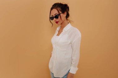 Koyu renk saçlı, kırmızı dudaklı, güneş gözlüklü, beyaz gömlekli, bej arkaplanda poz veren kendine güvenen şık bir bayan.