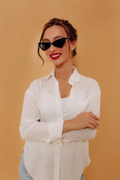 Αξιολάτρευτη όμορφη γυναίκα με σκούρα μαλλιά και κόκκινα χείλη που φορούν λευκό πουκάμισο σε μπεζ φόντο — Φωτογραφία Αρχείου