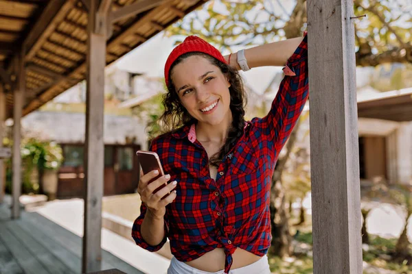 Excited charming lady in stretched shirt and red cap holding smartphone and smiling on summer terrace