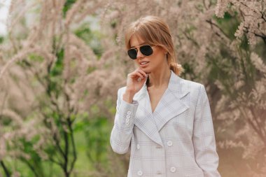 Yaz şehrinde poz veren muhteşem sarışın kadın. Çekici kız, açık renk takım elbise ve hoş aksesuarlar giyiyor, gülümsüyor ve bitkilerin arka planına dokunuyor. Yüksek kalite fotoğraf