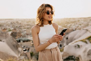 Günbatımında dağların arka planında akıllı telefon kullanarak beyaz tişört ve güneş gözlüğü takan modern kadın gezgin. Yüksek kalite fotoğraf