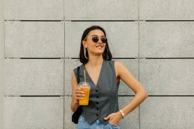 Gri yelek ve siyah güneş gözlüğü takmış şık modern model yaz içkisi içiyor ve gri duvarda poz veriyor. Yüksek kalite fotoğraf