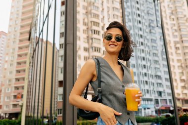 Koyu renk saçlı, gri yelek giyen, güneş gözlüklü, kameraya poz veren ve gülümseyen güzel bir kadın portresi. Yüksek kalite fotoğraf