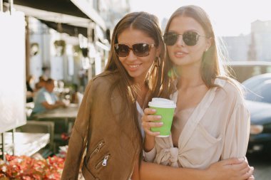 Dışarıda güneş gözlüğü ve bej bluz takan, güneş ışığında yürüyen ve kahve içen iki sevimli arkadaşın portresi var. Yüksek kalite fotoğraf