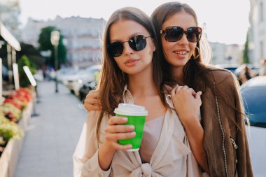 Bej bluz ve güneş gözlüğü takan şık kadınlar sokakta güneş ışığında sarılıp gülümsüyorlar. Yüksek kalite fotoğraf