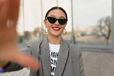 Koyu renk saçlı, kırmızı dudaklı, güneş gözlüğü takan ve gri ceketli, şehirde selfie çeken çekici bir kız.