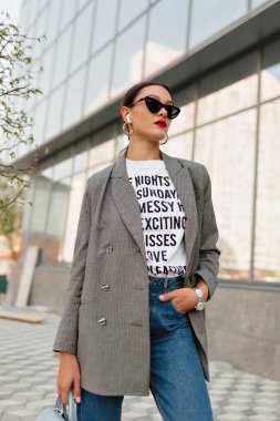 Gri ceketli, güneş gözlüklü ve güneşli bir günde sokakta poz veren hoş bir bayan.