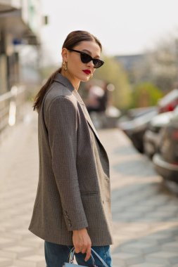 Arka planda gün ışığında yürüyen zarif bir kızın portresi. Gri ceket ve kot pantolon giyiyor. Kameranın önünde gülümsüyor..