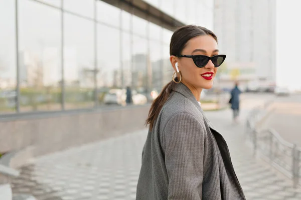 Wanita cantik dengan rambut hitam dan bibir merah memakai kacamata hitam dan jaket berjalan di alun-alun kota di musim panas yang hangat — Stok Foto