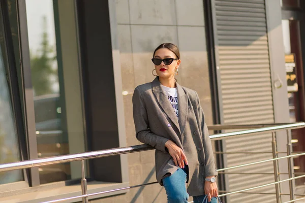 Mujer joven con estilo en traje formal posando en la calle en la ciudad. Elegante morena en gafas de sol disfrutando de las vistas de la ciudad por la mañana. — Foto de Stock