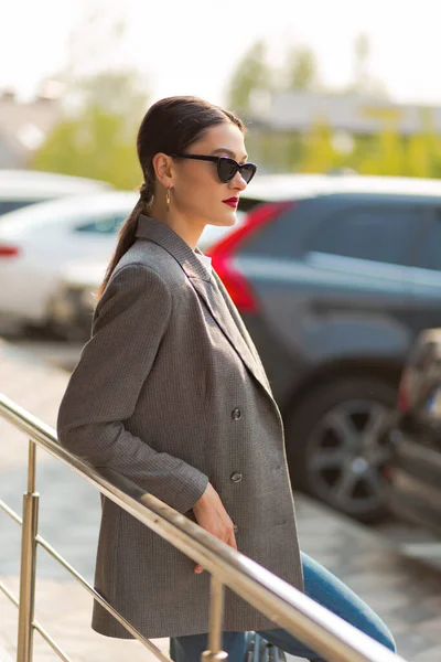 Retrato de una mujer de negocios con estilo en una chaqueta gris en las escaleras al aire libre. Lleva gafas de sol y se ve recta. — Foto de Stock