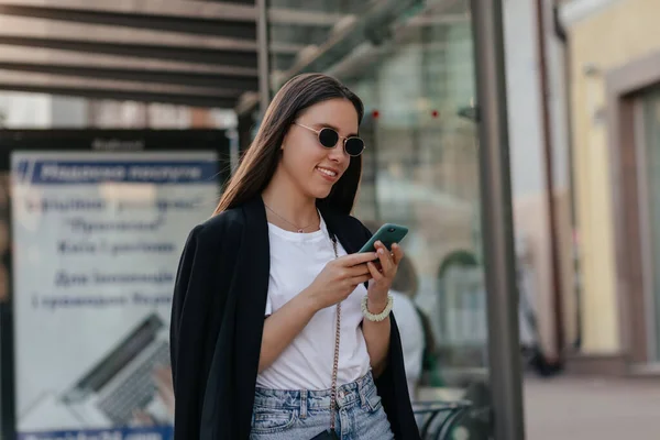 Pretty woman in black jacket and shite shirt holds phone outside. European brunette lady walks and looks away outdoors. — Stok Foto