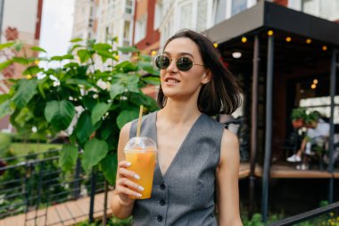 Kısa saçlı, güneş gözlüklü, şehir kafeteryasının üstünde meyve suyuyla duran sevimli bir kadın.
