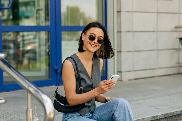 Cukup tersenyum gadis dengan gaya rambut pendek mengenakan rompi abu-abu dan jeans duduk di tangga dan menggunakan smartphone dengan senyum bahagia — Stok Foto